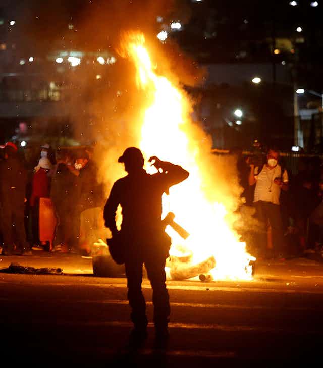 A policeman standing in front of a raging fire as a group of people block a road.