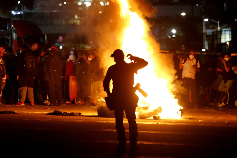 A policeman standing in front of a raging fire as a group of people block a road.