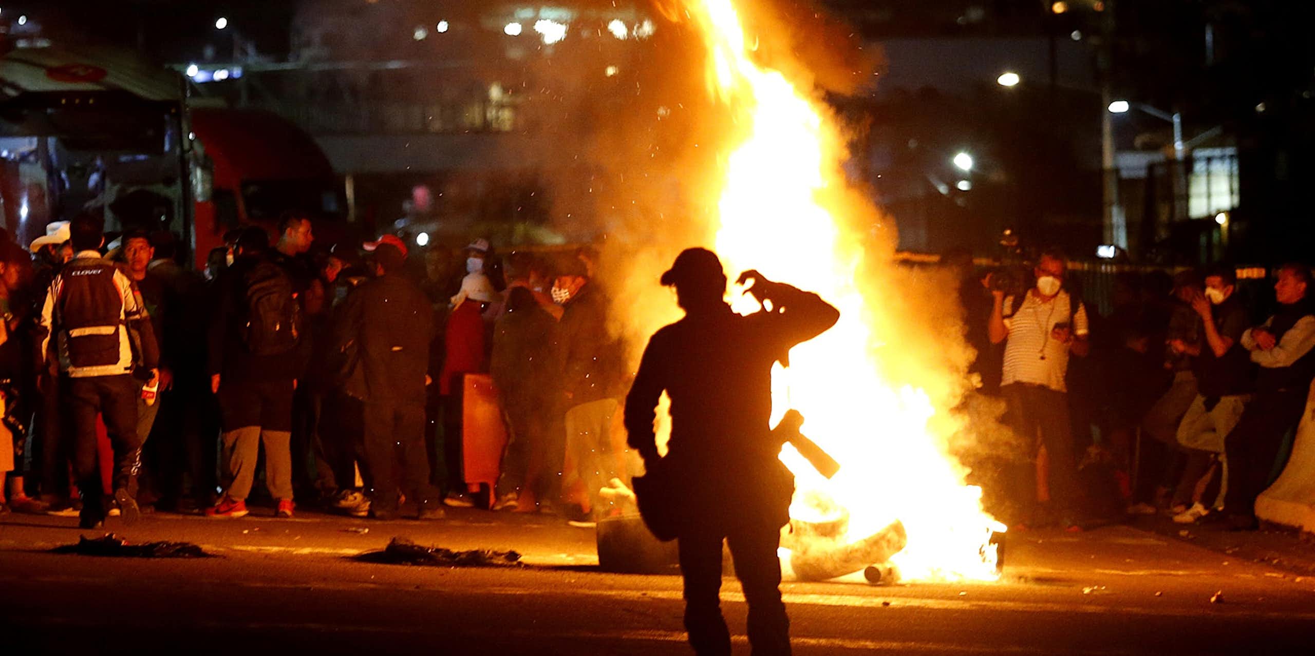 A policeman standing in front of a raging fire as a group of people block a road.