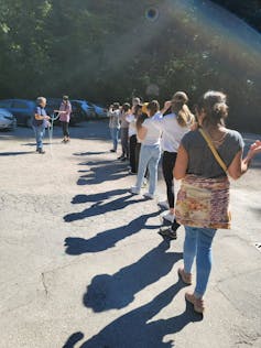 Nine people outside. Many people stand in a line while one person is blindfolded and holding a string and others are lifting their blindfolds.