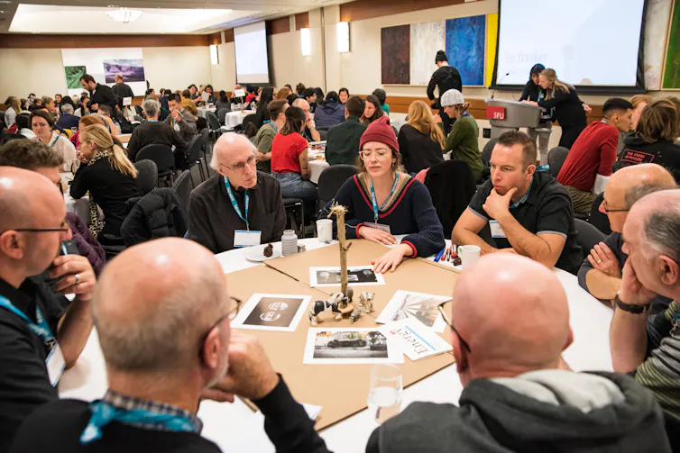 People at a roundtable in discussion and a full room of other roundtables is seen in the background.
