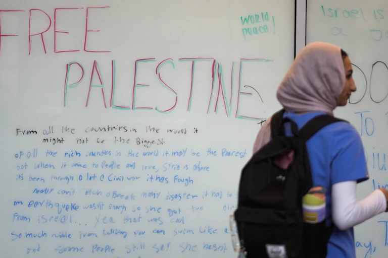 The words 'world peace' and 'free Palestine' seen on a white board as a woman in hijab and knapsack walks past.