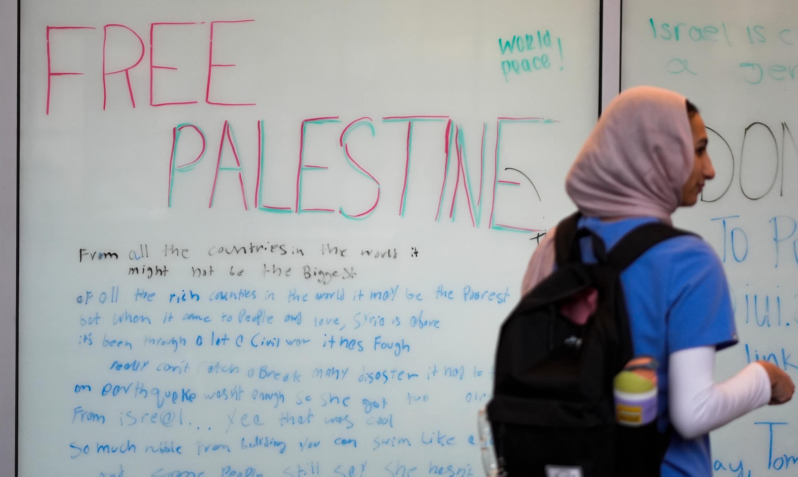 The words 'world peace' and 'free Palestine' seen on a white board as a woman in hijab and knapsack walks past.