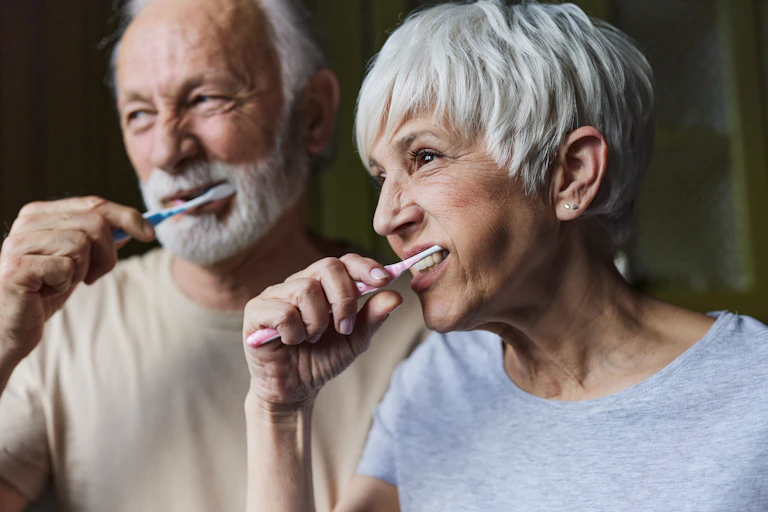 Older man and older woman brush their teeth.