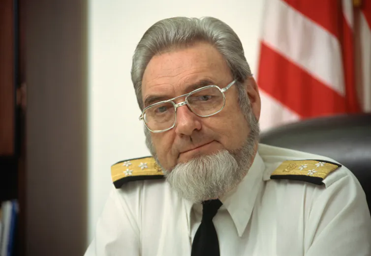 Photo of a man in a military-looking uniform, wearing a bushy gray goatee and large wire-rimmed glasses, with an American flag in the backgorund.