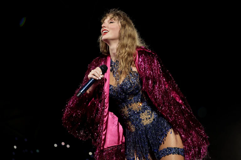 A blond woman smiles as she sings in a sparkly purple robe and blue bodysuit.
