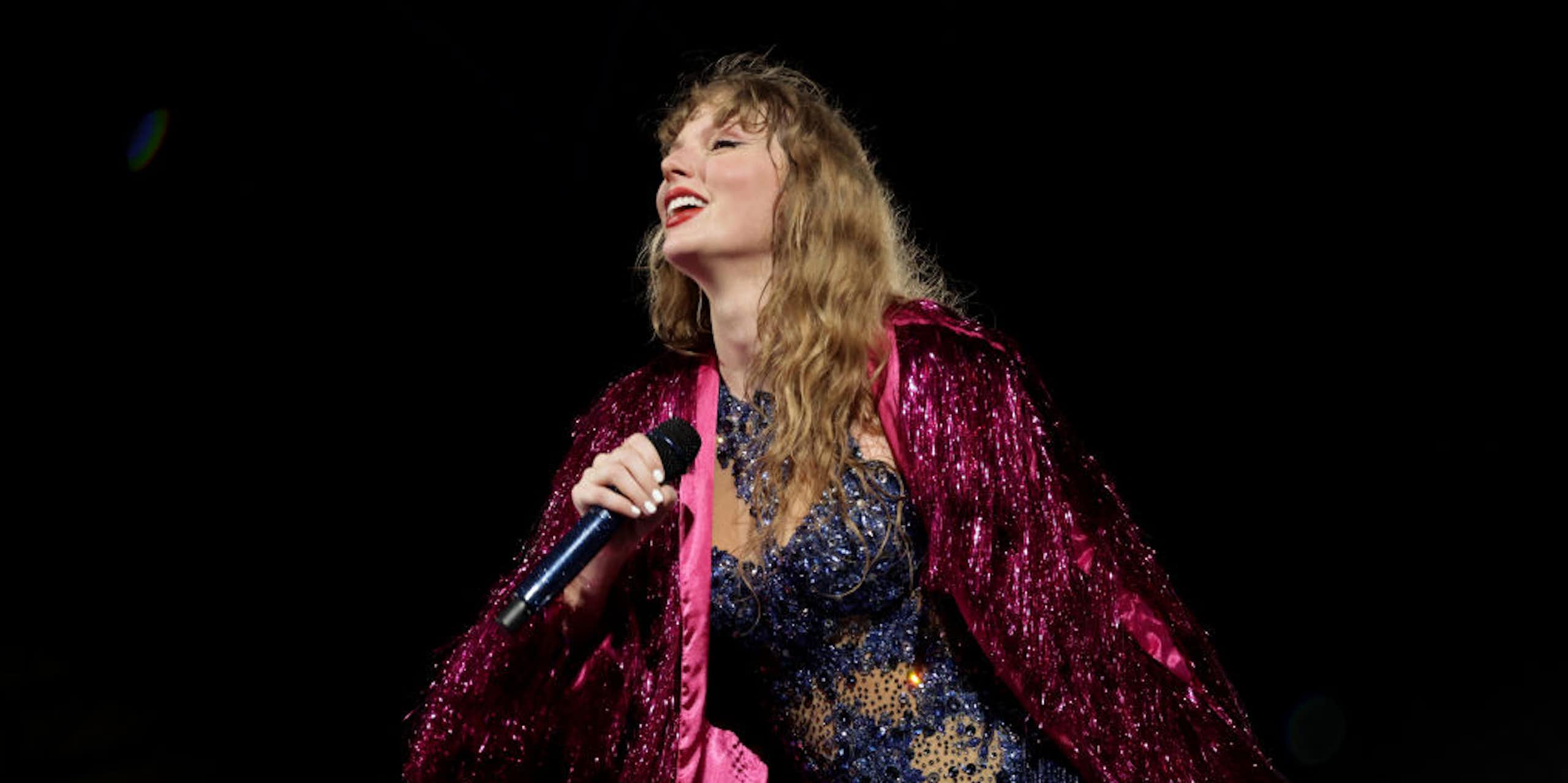 A blond woman smiles as she sings in a sparkly purple robe and blue bodysuit.