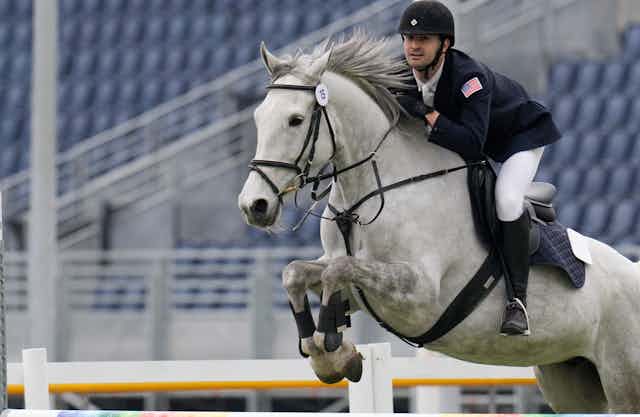 A horseback rider leans forward in the saddle as a gray horse jumps over an obstacle