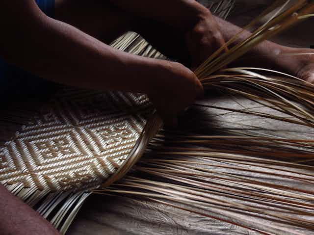 Foto mostra as mãos de um indígena tecendo fibras vegetais e criando um padrão geométrico de 2 cores, bege e marrom