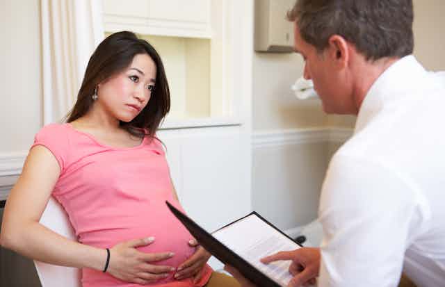 A pregnant woman in a pink shirt with her hands on her belly, and a health-care worker seen from behind