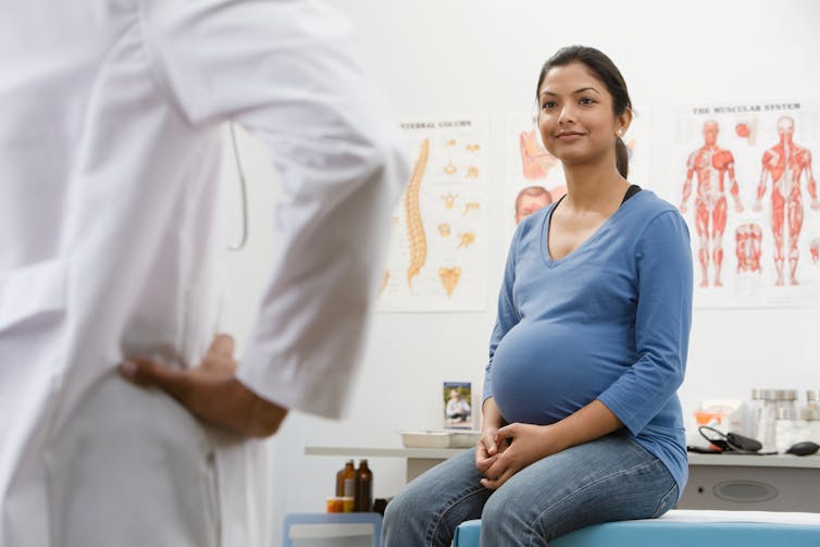 A pregnant woman in a doctor's office with a health-care worker in a white coat seen from behind