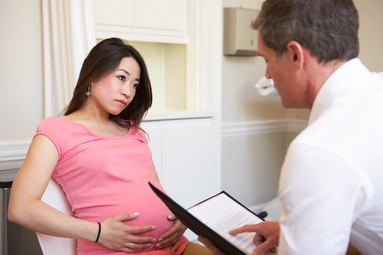 Une femme enceinte vêtue d'une chemise rose, les mains sur le ventre, et un médecin vu de dos.