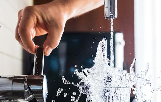 A hand turns on a faucet and water splashes form a full glass.
