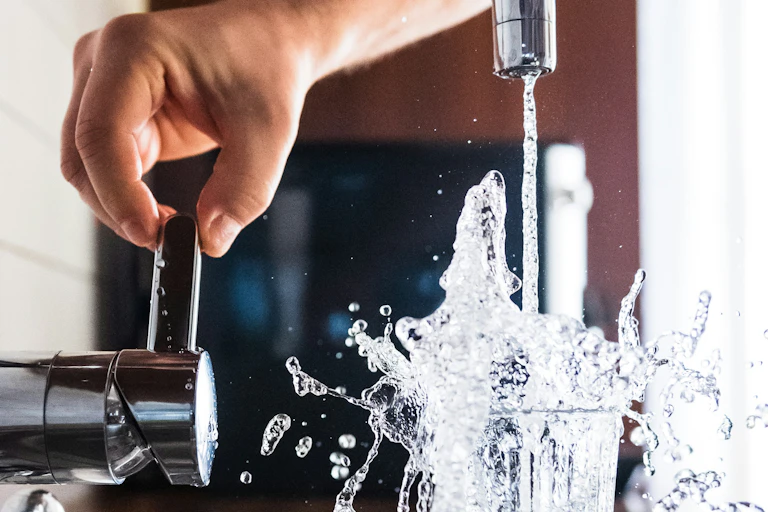 A hand turns on a faucet and water splashes form a full glass.