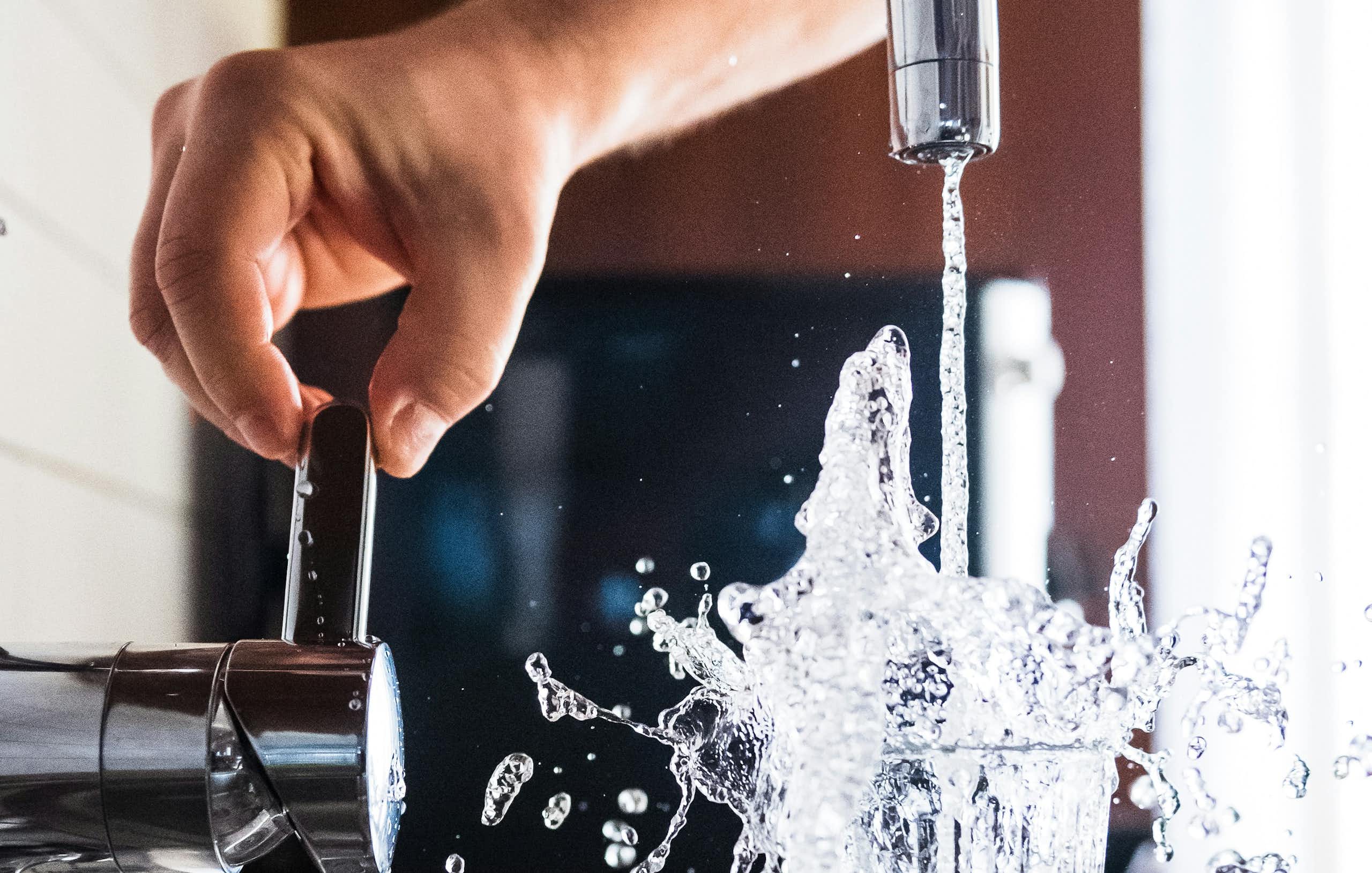 A hand turns on a faucet and water splashes form a full glass.