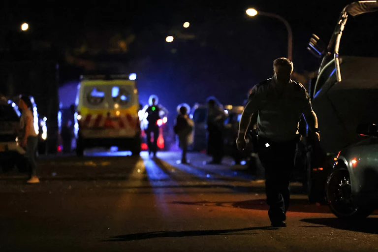 A police officer walks down a dark street, lit by emergency lights