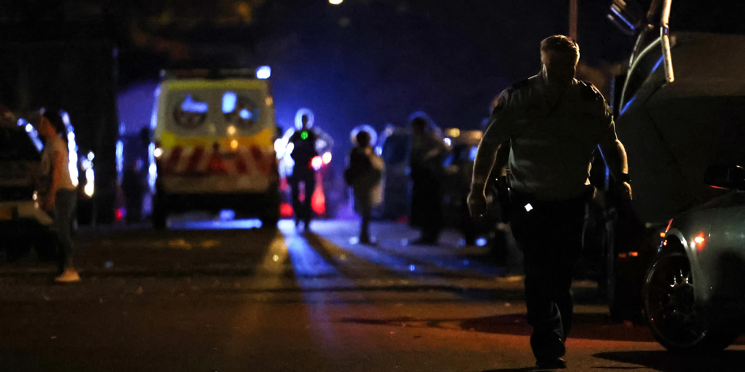 A police officer walks down a dark street, lit by emergency lights