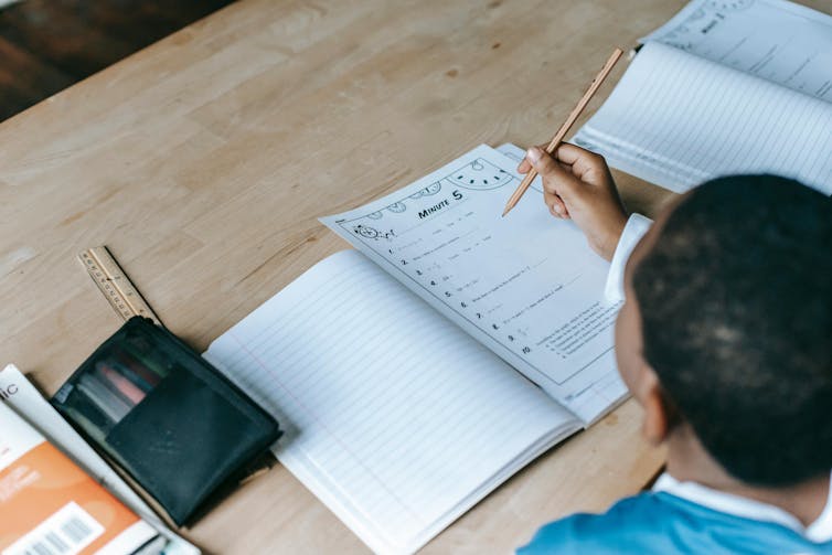 A student works on a maths worksheet, with papers and rulers on a desk.