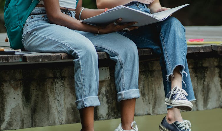 Two young people in jeans and sneakers sit on a low wall, with a folder.