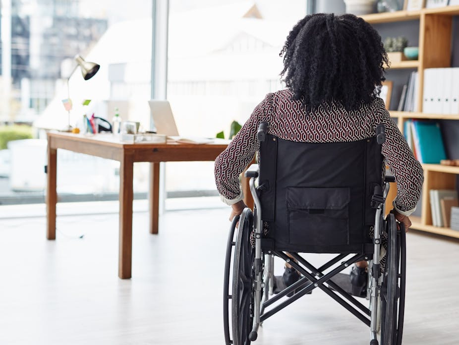 Rearview photo of a woman sitting in her wheelchair, in an office with bookshelves and a desk in the background