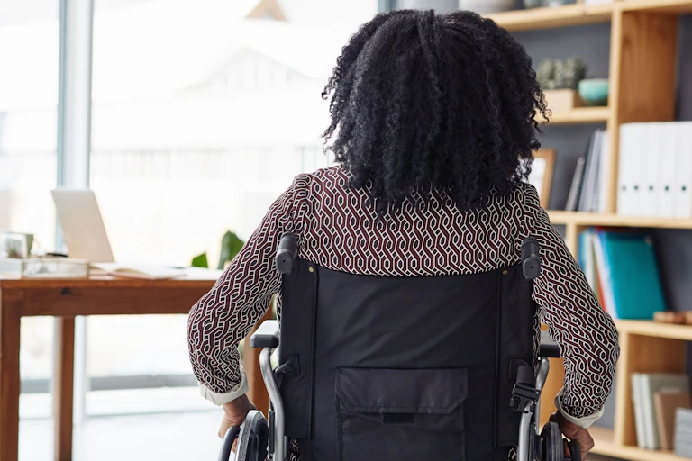 Rearview photo of a woman sitting in her wheelchair, in an office with bookshelves and a desk in the background