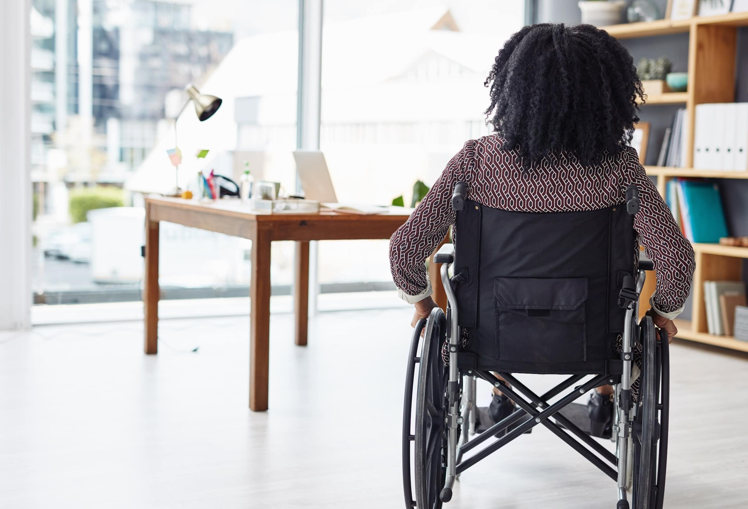 Rearview photo of a woman sitting in her wheelchair, in an office with bookshelves and a desk in the background