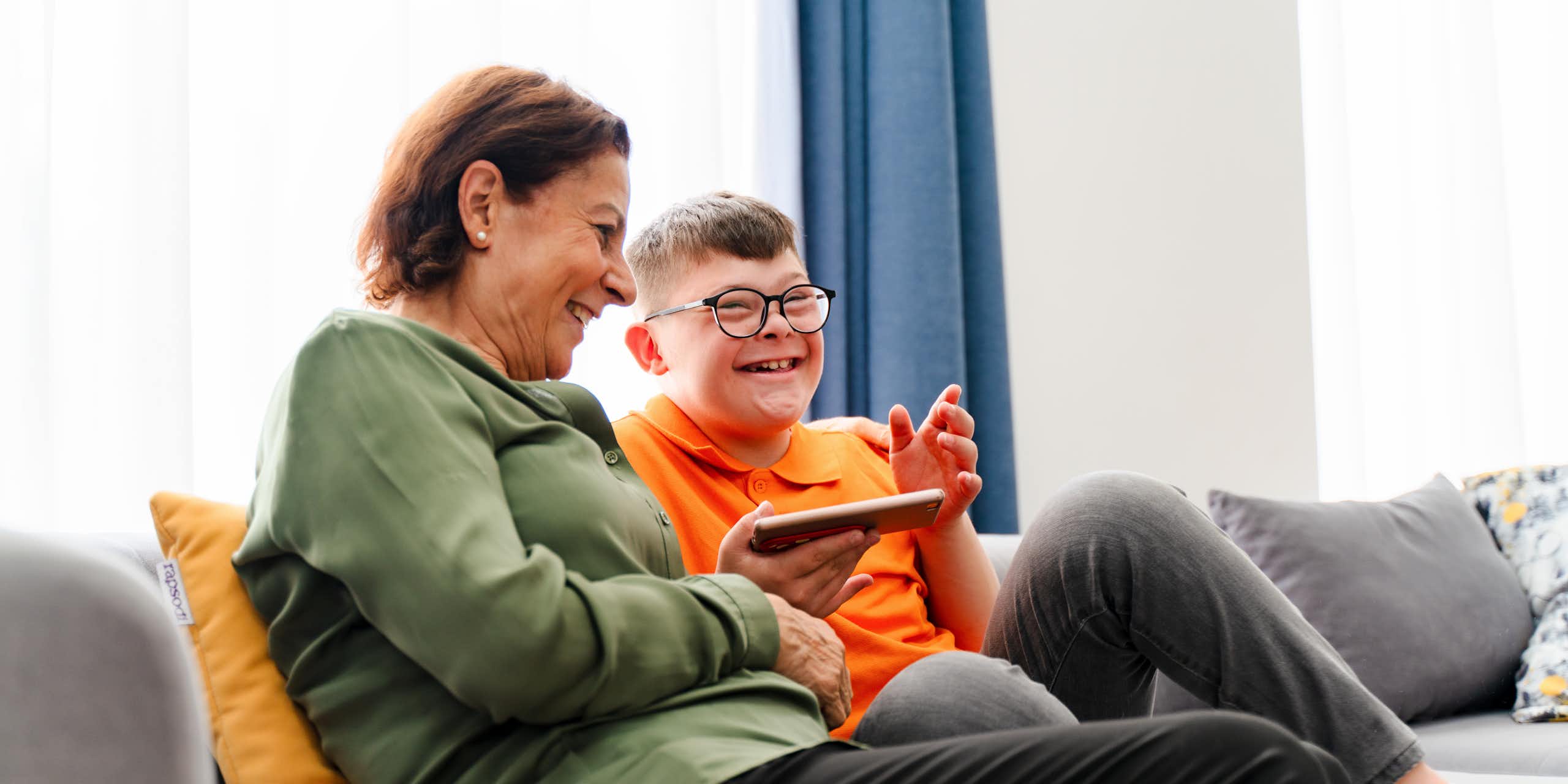 Woman and younger man with down syndrome sit together on couch. Both are laughing