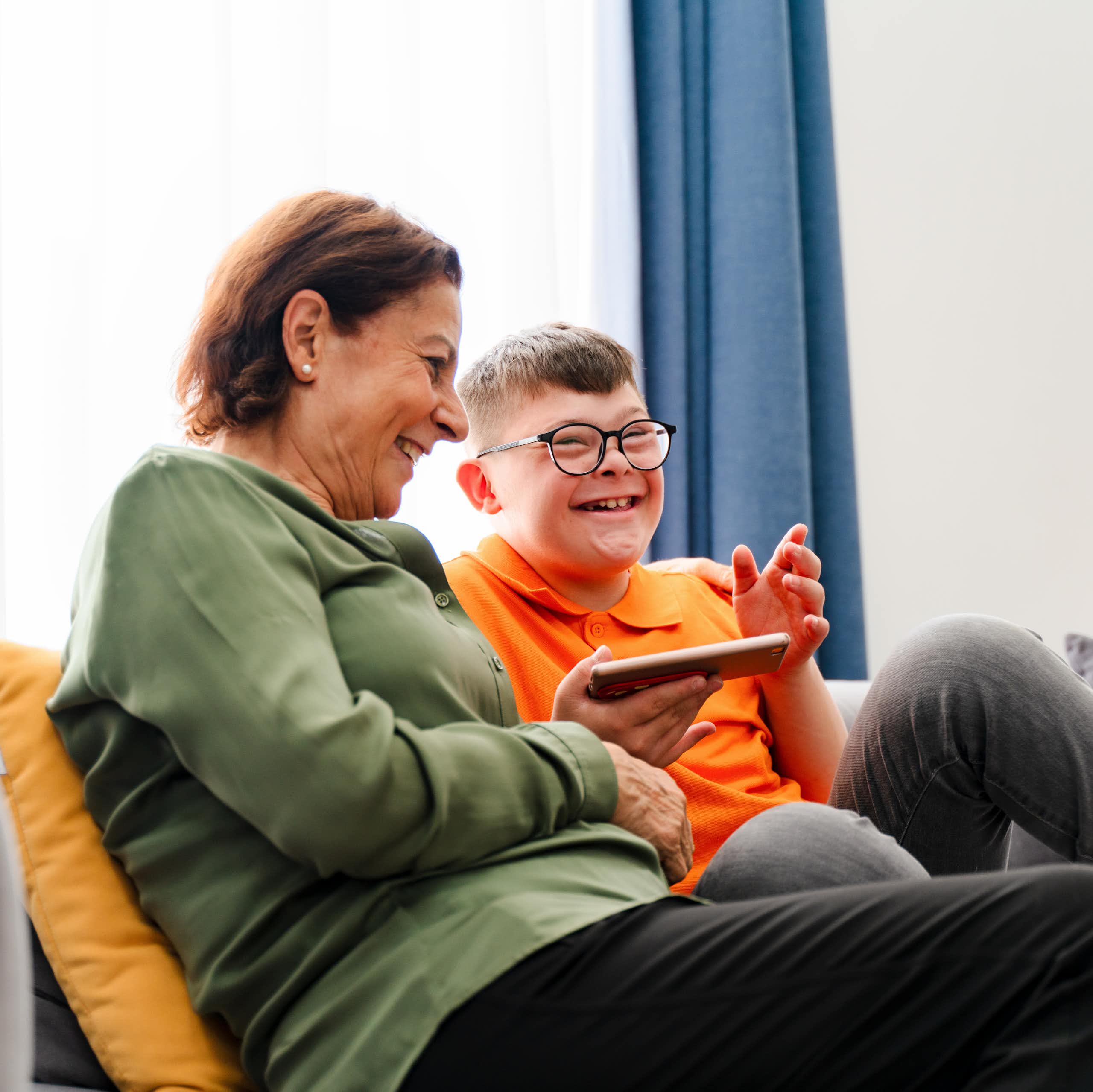 Woman and younger man with down syndrome sit together on couch. Both are laughing