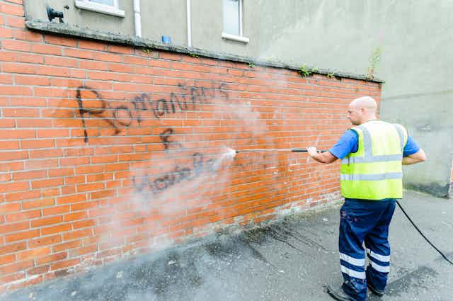 A council worker power washes hateful graffiti off a red brick wall. 