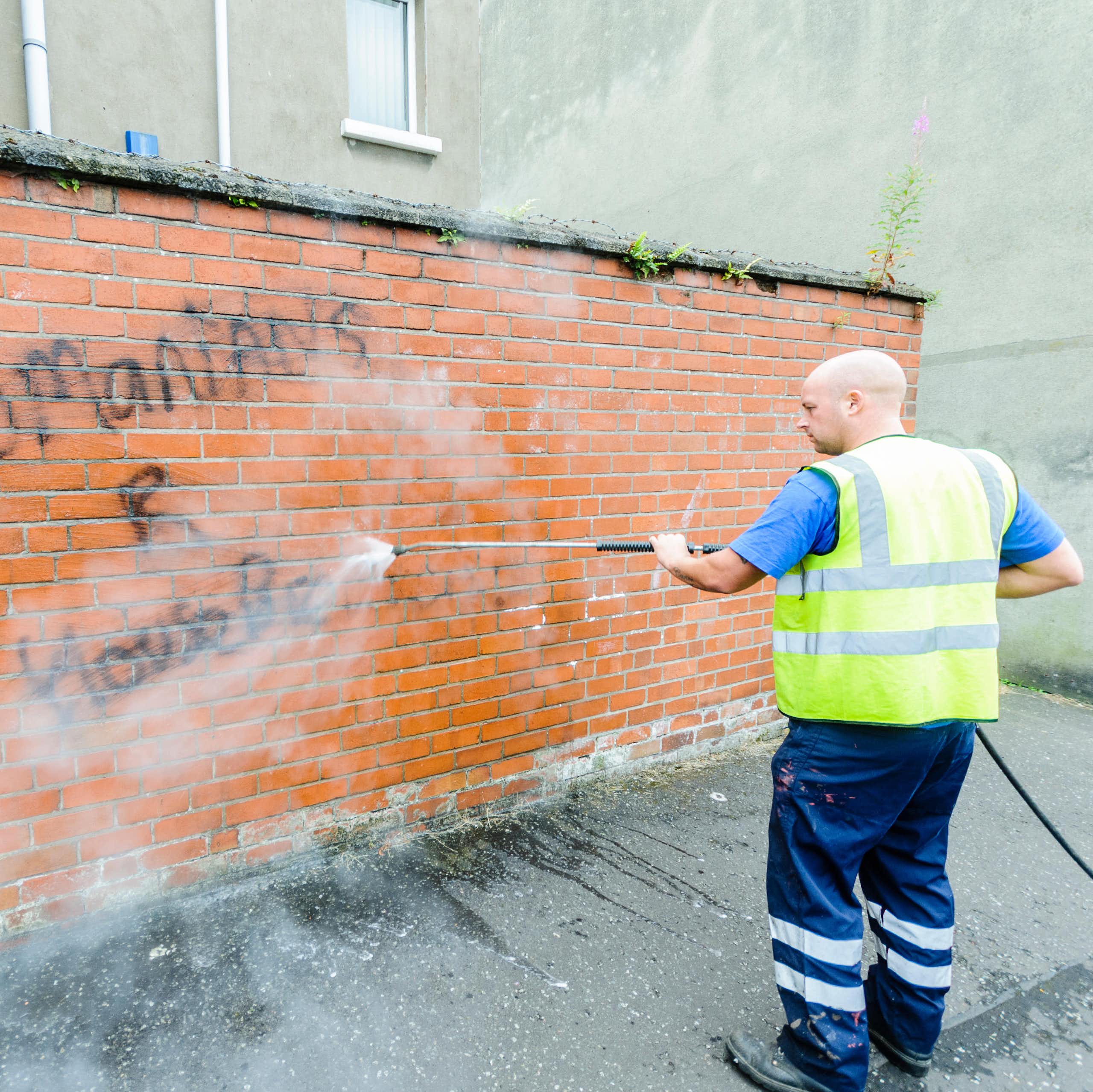 A council worker power washes hateful graffiti off a red brick wall. 