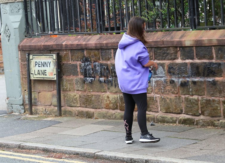 A woman wearing lilac stands in front of a wall on Penny Lane in Liverpool.