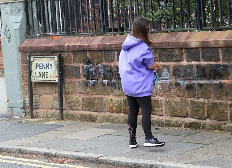 A woman wearing lilac stands in front of a wall on Penny Lane in Liverpool.