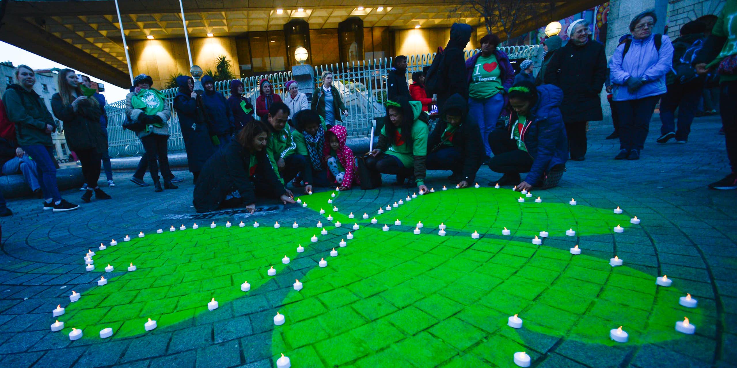 A green shamrock is painted on stone pavement surrounded by people in 'I heart Ireland' T-shirts.