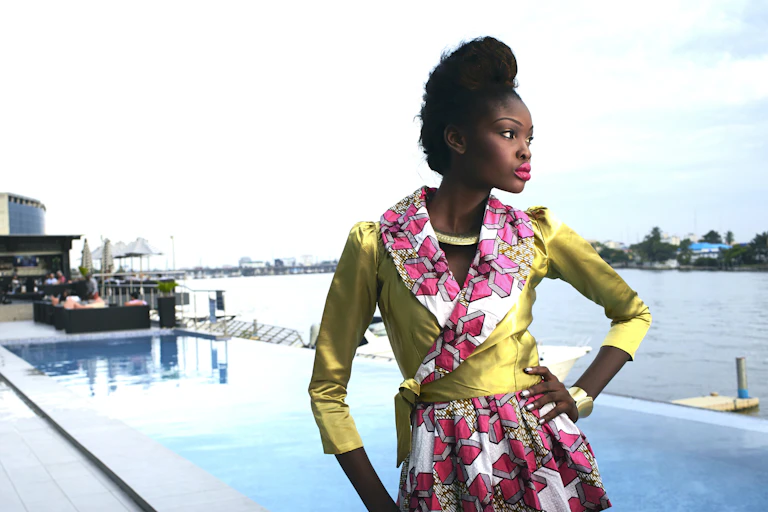 A young woman stands at a waterfront pool with an arm on her hip, posing in an outfit of shiny green with pink African fabric contrasting it on the collar and skirt.