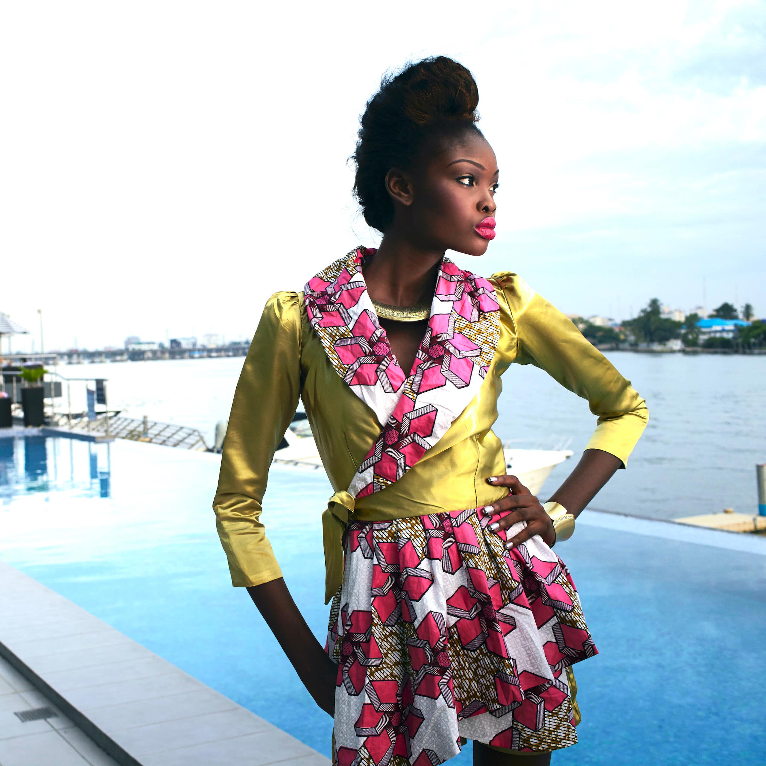  A young woman stands at a waterfront pool with an arm on her hip, posing in an outfit of shiny green with pink African fabric contrasting it on the collar and skirt. 