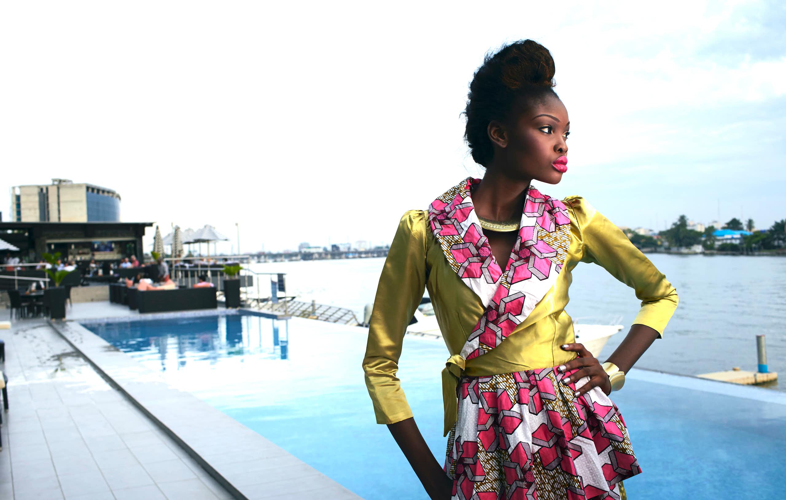  A young woman stands at a waterfront pool with an arm on her hip, posing in an outfit of shiny green with pink African fabric contrasting it on the collar and skirt. 