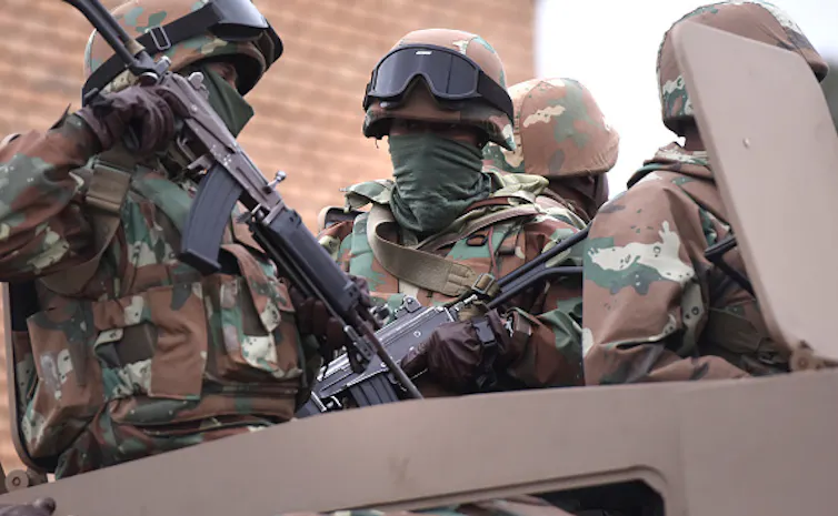 Soldiers bearing rifles travel in an armoured vehicle.