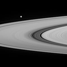 Image en noir et blanc d’une portion des anneaux de Saturne, avec la lune Mimas en arrière plan. On distingue la division de Cassini, une portion plus sombre parmi les anneaux.
