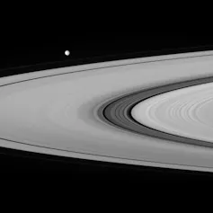Image en noir et blanc d’une portion des anneaux de Saturne, avec la lune Mimas en arrière plan. On distingue la division de Cassini, une portion plus sombre parmi les anneaux.