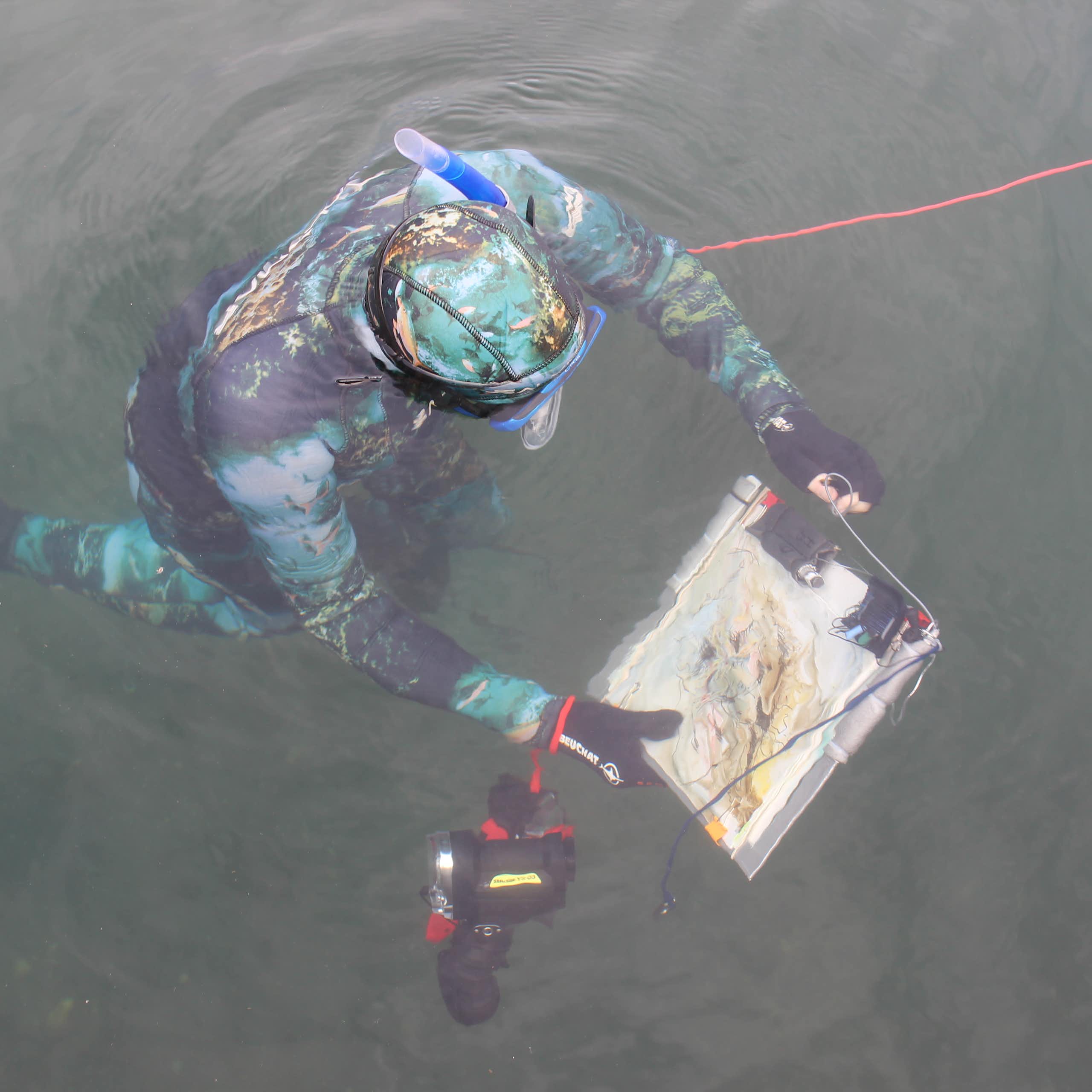 looking down at snorkellor in camoflage coloured wetsuit, holding clipboard with white paper and painting on, dark blue-grey sea surrounding them
