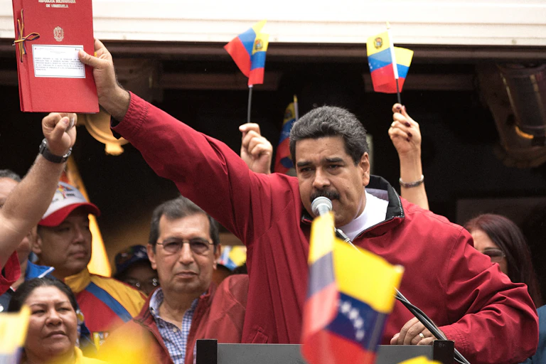 Venezuelan president Nicolas Maduro holding a red book with Venezuela flags waving around him.