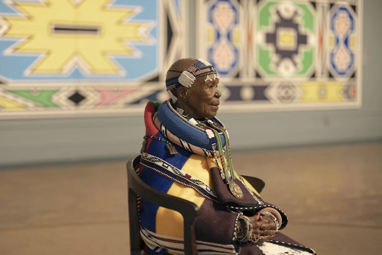 An elderly woman sits on a chair in an art gallery full of bright murals. She wears intricate traditional Ndebele clothing with beaded head wrap and outsized beaded necklaces as well as a blanket in brown, yellow, navy and red.