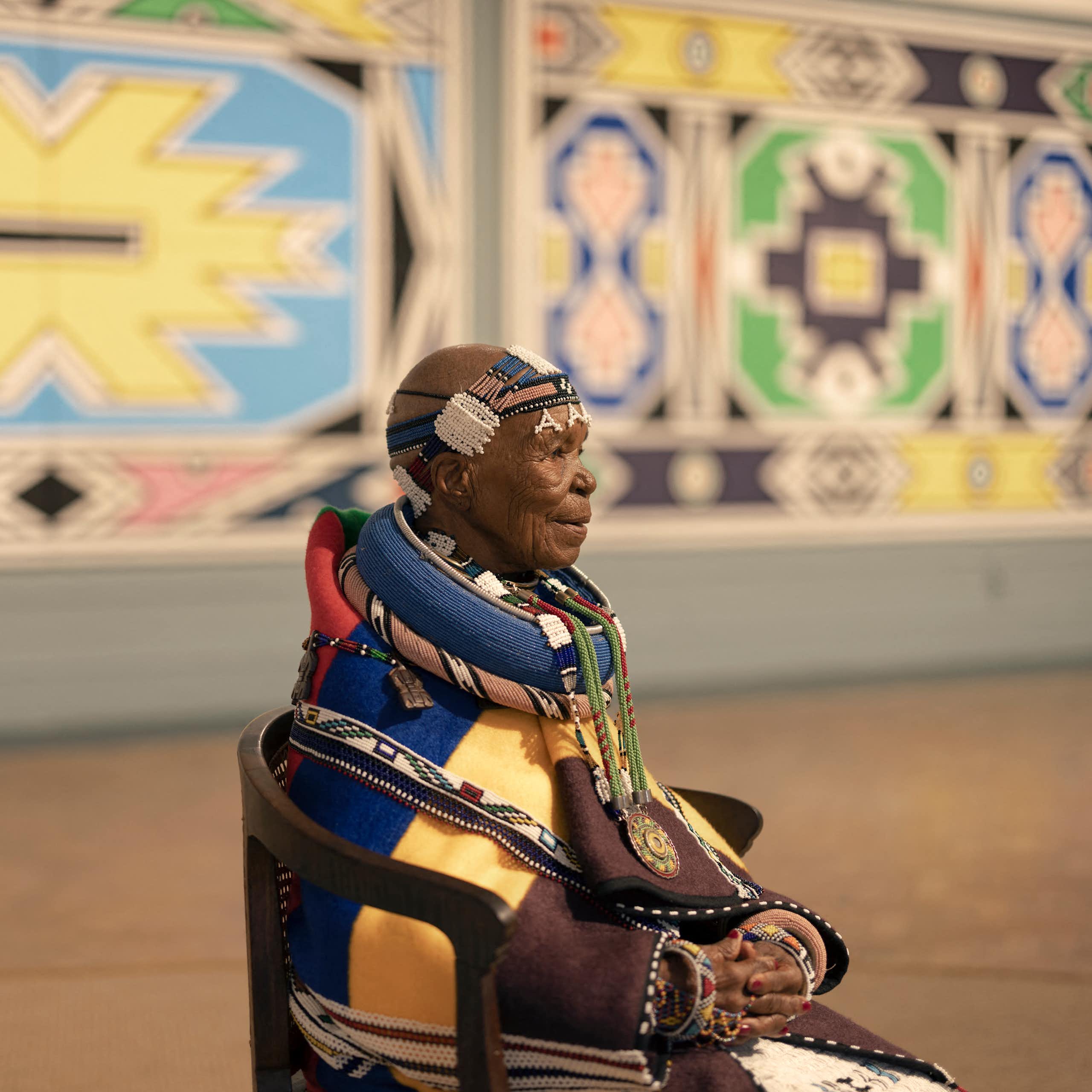 An elderly woman sits on a chair in an art gallery full of bright murals. She wears intricate traditional Ndebele clothing with beaded head wrap and outsized beaded necklaces as well as a blanket in brown, yellow, navy and red.