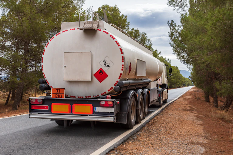 A large semi truck with eight wheels and a large cylindrical tank with a red flame sticker on the back drives down a road.