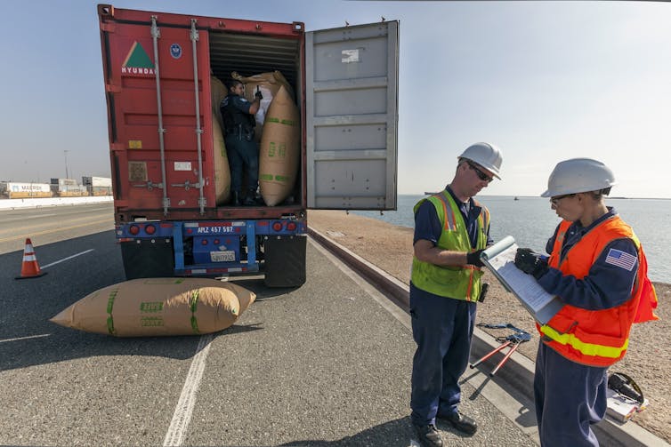 Two men in light-colored vests and hard hats talk while standing in front of a semi-truck with the bed open and several large sacks visible.