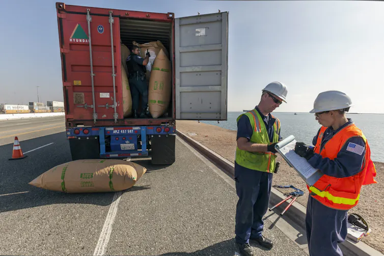 Two men wearing bright vests and hard hats talk while standing in front of a semi truck with the back open and several large sacks visible.