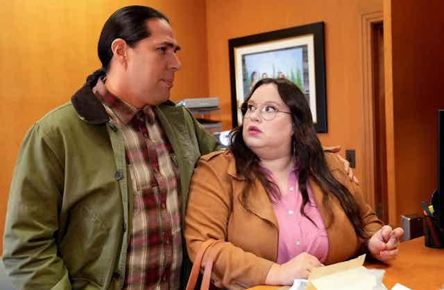 Man wearing olive green jacket side hugs a woman wearing a brown-colored trench coat.