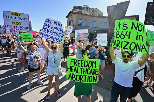 People stand in the streets and hold signs that say, 'It's not 1864 in Arizona,' 'Freedom healthcare abortion,' and 'November November November November.'