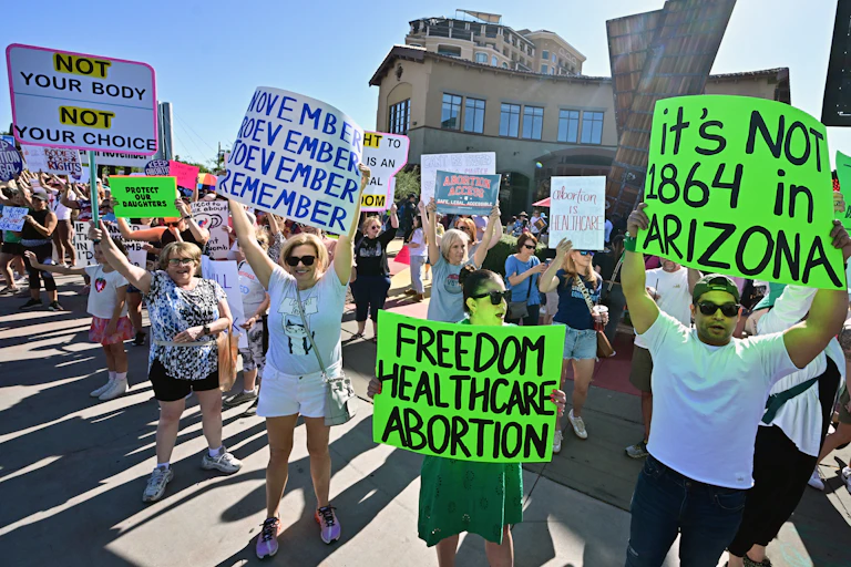 People stand in the streets and hold signs that say, 'It's not 1864 in Arizona,' 'Freedom healthcare abortion,' and 'November November November November.'