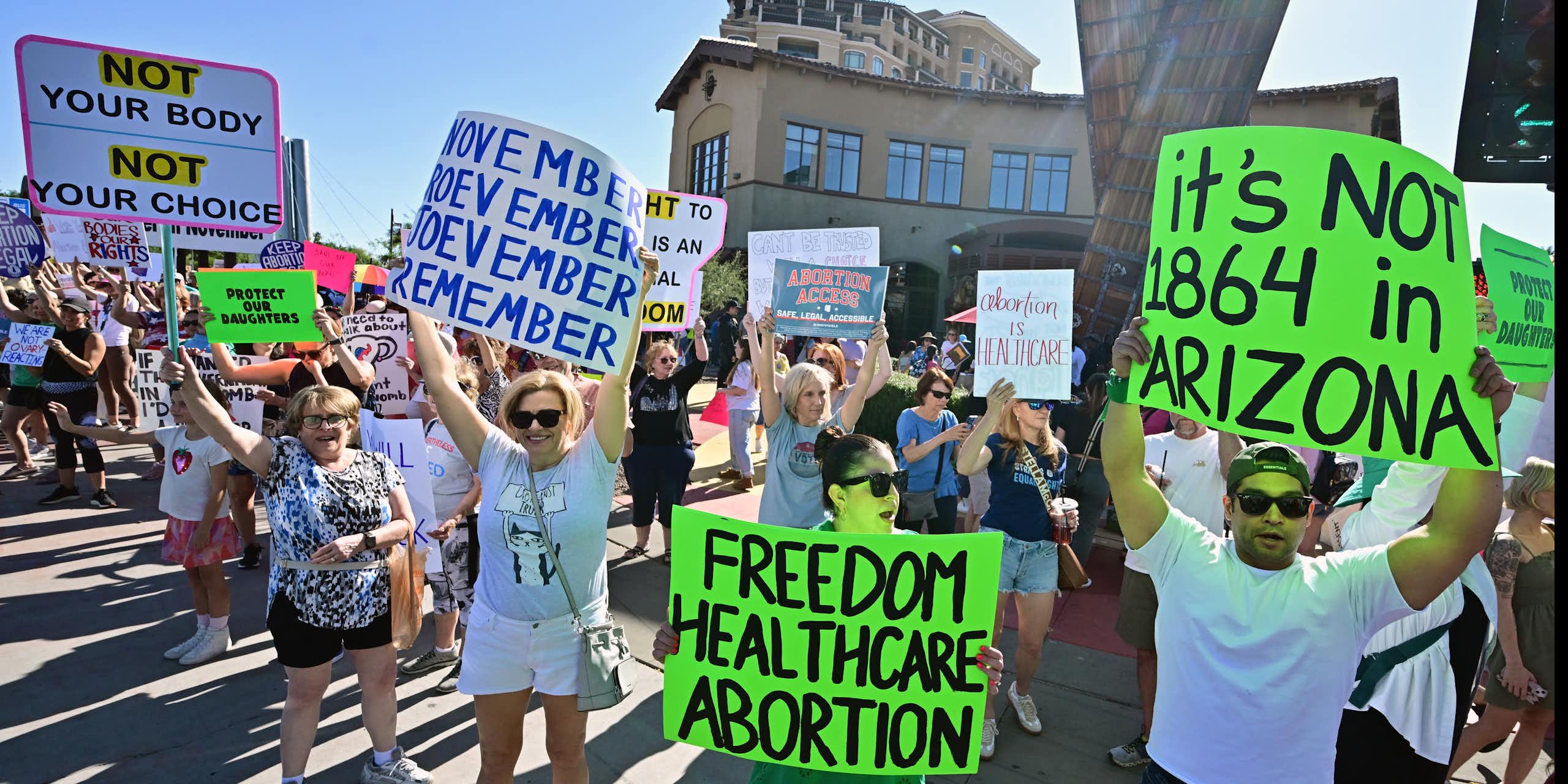 People stand in the streets and hold signs that say, 'It's not 1864 in Arizona,' 'Freedom healthcare abortion,' and 'November November November November.'